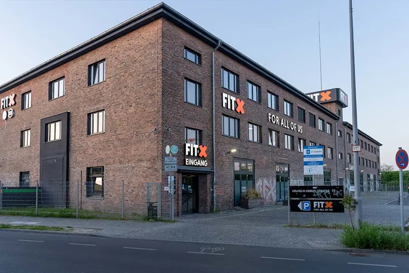 Bild: Außenansicht eines FitX Fitnessstudios. Ein großes FitX-Logo mit orangefarbenem X ist zu sehen. Das Gebäude befindet sich in einem urbanen Umfeld.