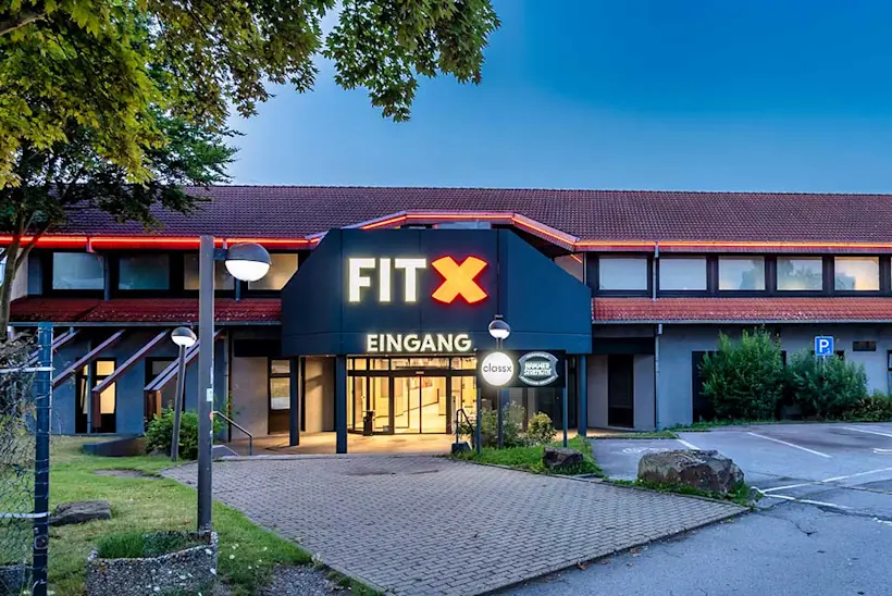 Bild: Außenansicht eines FitX Fitnessstudios. Ein großes FitX-Logo mit orangefarbenem X ist zu sehen. Das Gebäude befindet sich in einem urbanen Umfeld.