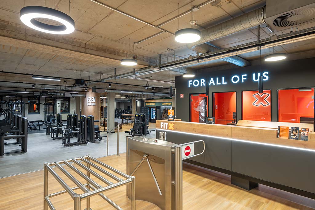 Empfangsbereich des FitX Fitnessstudios Essen-Bergerhausen mit einer holzverkleideten Theke, einem Drehkreuz am Eingang und dem leuchtenden Schriftzug „FOR ALL OF US“ an der Wand.