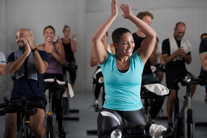 Bild: Teilnehmende im tour-de-x-Kurs sitzen auf Indoor-Bikes, lachen und klatschen.