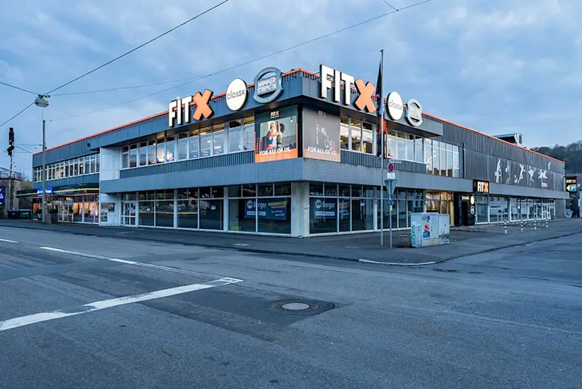 Bild: Außenansicht eines FitX Fitnessstudios. Ein großes FitX-Logo mit orangefarbenem X ist zu sehen. Das Gebäude befindet sich in einem urbanen Umfeld.