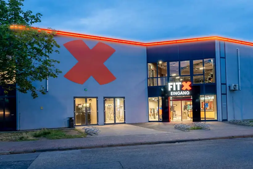 Bild: Außenansicht eines FitX Fitnessstudios. Ein großes FitX-Logo mit orangefarbenem X ist zu sehen. Das Gebäude befindet sich in einem urbanen Umfeld.