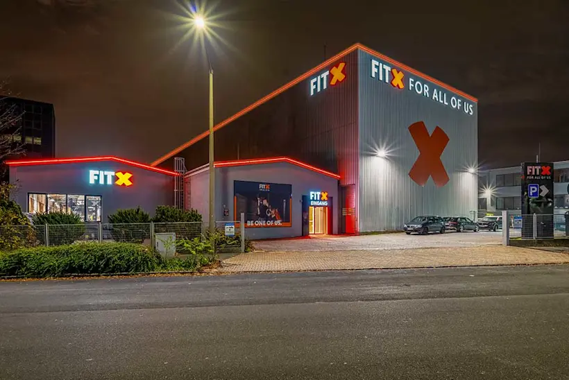 Bild: Außenansicht des FitX-Studios in Fürth am Abend. Das Gebäude hat zwei einstöckige Gebäude. Rechts befindet sich ein großes, Lagerhallenartiges Gebäude. Auf den Außenfassaden leuchtet das FitX-Logo in Form unterschiedlicher Leuchtschilder. Auf dem mehrstöckigen Teil des Gebäudes ist ein großes, orangenes X.