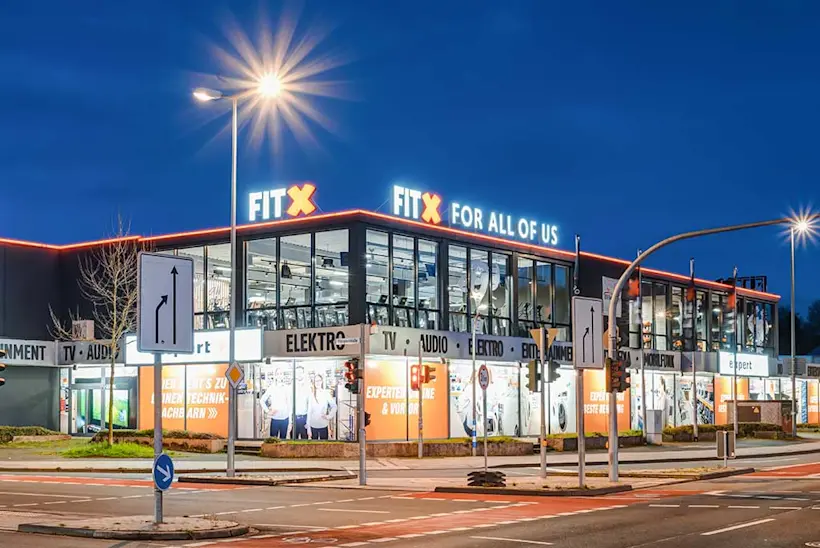 Bild: Außenansicht eines FitX Fitnessstudios. Ein großes FitX-Logo mit orangefarbenem X ist zu sehen. Das Gebäude befindet sich in einem urbanen Umfeld.