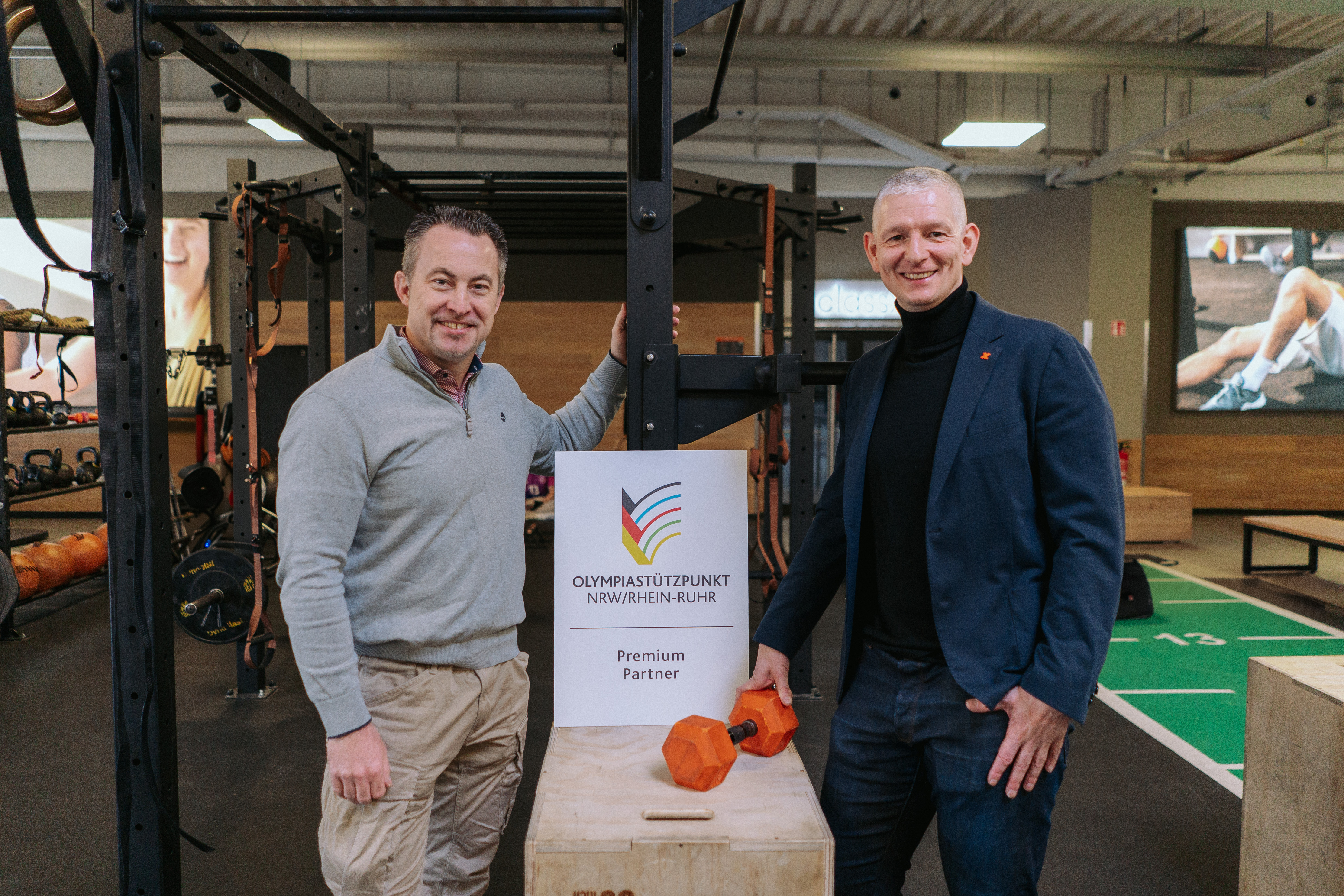 Bild: Das Bild zeigt FitX-CEO Markus Vancraeyenest (rechts im Bild) und Leiter des Olympiastützpunkt NRW/Rhein-Ruhr Volker Lauer (links im Bild). In der Mitte des Bildes steht ein Schild mit dem Logo des Olympiastützpunkt mit der Unterschrift 
