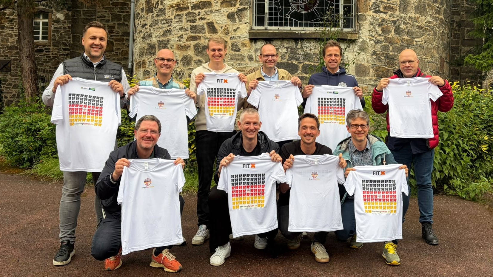 Bild: Zehn Männer stehen nebeneinander und blicken in die Kamera. Jeder hält ein bedrucktes Shirt in den Händen und zeigt es in die Kamera.