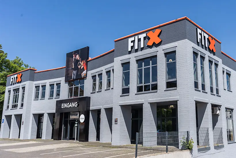 Bild: Außenansicht eines FitX Fitnessstudios. Ein großes FitX-Logo mit orangefarbenem X ist zu sehen. Das Gebäude befindet sich in einem urbanen Umfeld.