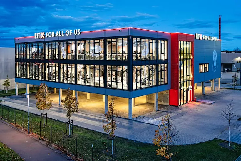 Bild: Außenansicht eines FitX Fitnessstudios. Ein großes FitX-Logo mit orangefarbenem X ist zu sehen. Das Gebäude befindet sich in einem urbanen Umfeld.