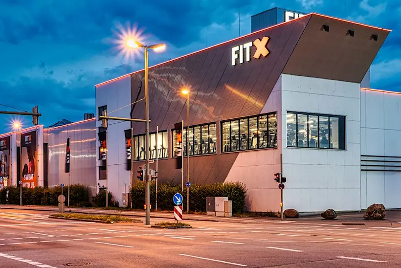 Bild: Außenansicht eines FitX Fitnessstudios. Ein großes FitX-Logo mit orangefarbenem X ist zu sehen. Das Gebäude befindet sich in einem urbanen Umfeld.