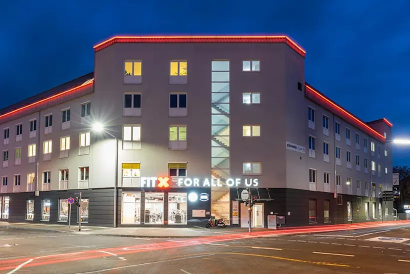 Bild: Außenansicht eines FitX Fitnessstudios. Ein großes FitX-Logo mit orangefarbenem X ist zu sehen. Das Gebäude befindet sich in einem urbanen Umfeld.