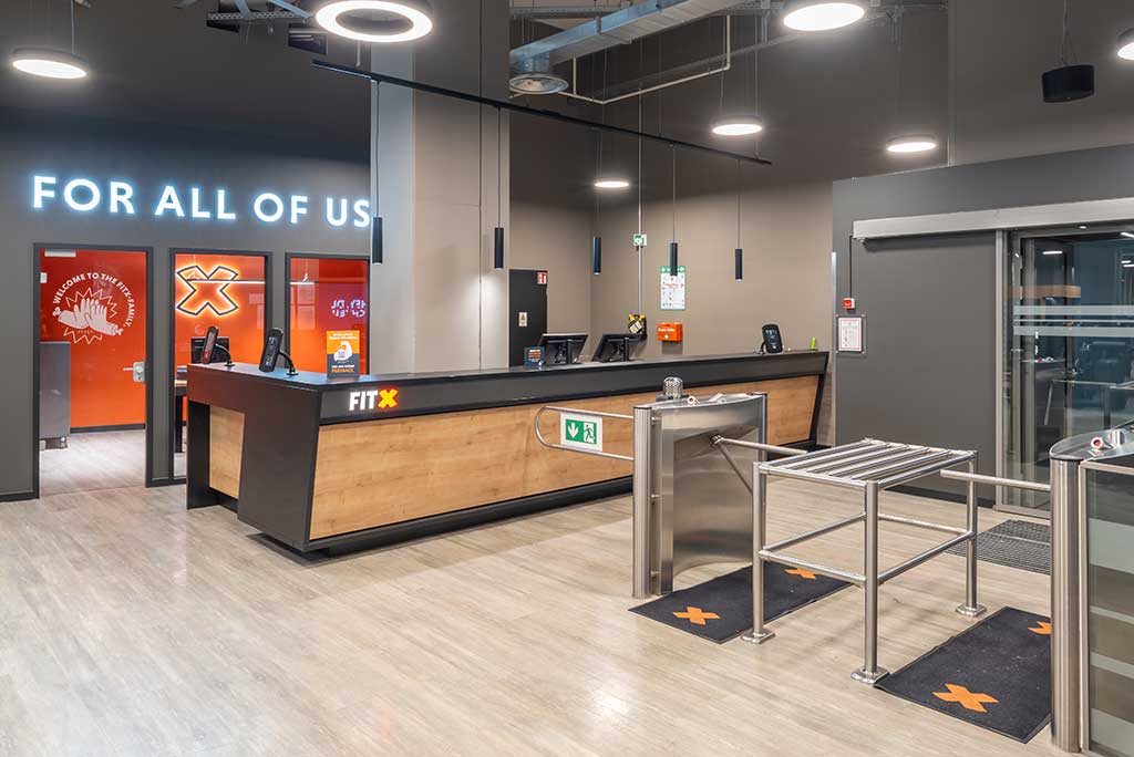 Empfangsbereich des FitX Fitnessstudios Gießen-Innenstadt mit einer holzverkleideten Theke, einem Drehkreuz am Eingang und dem leuchtenden Schriftzug „FOR ALL OF US“ an der Wand.