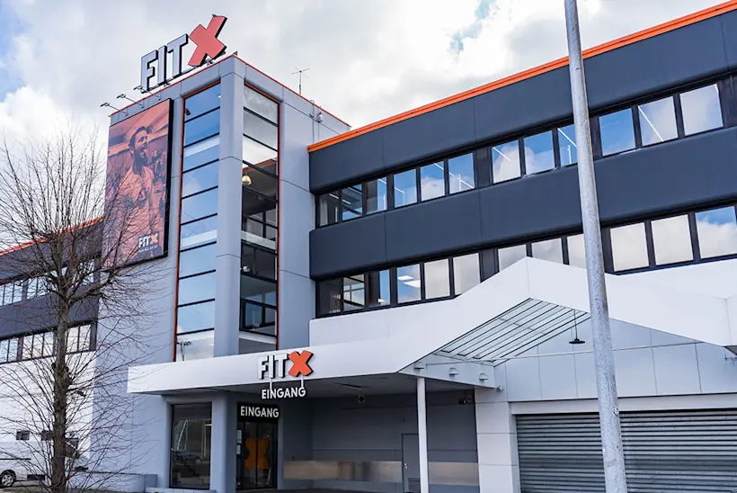 Bild: Außenansicht eines FitX Fitnessstudios. Ein großes FitX-Logo mit orangefarbenem X ist zu sehen. Das Gebäude befindet sich in einem urbanen Umfeld.