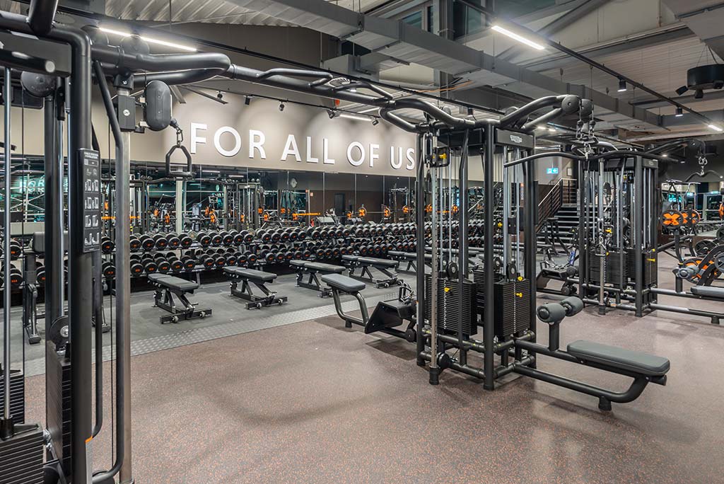 Weitläufiger Freihantelbereich mit verschiedenen Kraftgeräten im FitX Fitnessstudio Neuss-Innenstadt. An der Seite sind Kurzhanteln vor einer Spiegelwand aufgereiht. An der oberen Wand prangt der Schriftzug „FOR ALL OF US“.