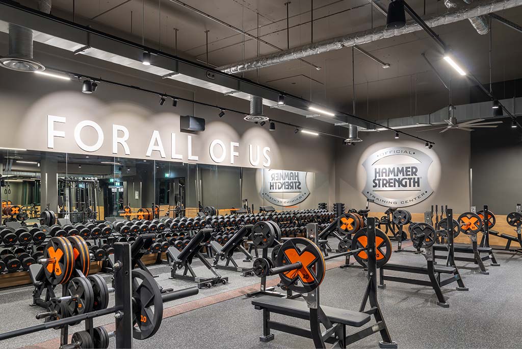 Weitläufiger Freihantelbereich mit verschiedenen Kraftgeräten im FitX Fitnessstudio Düsseldorf-Derendorf. An der Seite sind Kurzhanteln vor einer Spiegelwand aufgereiht. An der oberen Wand prangt der Schriftzug „FOR ALL OF US“.