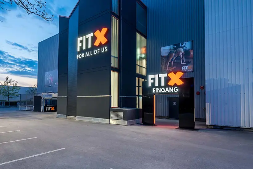 Bild: Außenansicht eines FitX Fitnessstudios. Ein großes FitX-Logo mit orangefarbenem X ist zu sehen. Das Gebäude befindet sich in einem urbanen Umfeld.