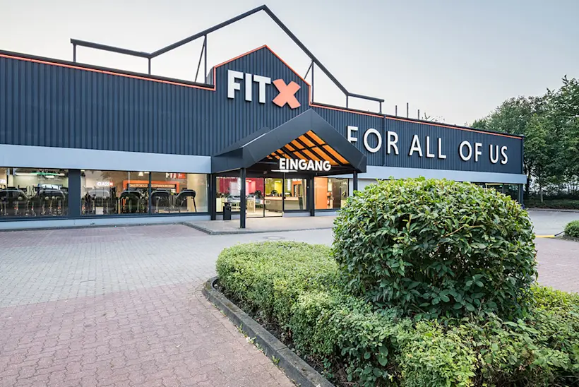 Bild: Außenansicht eines FitX Fitnessstudios. Ein großes FitX-Logo mit orangefarbenem X ist zu sehen. Das Gebäude befindet sich in einem urbanen Umfeld.
