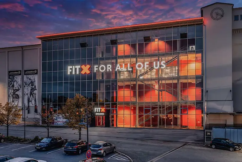 Bild: Außenansicht eines FitX Fitnessstudios. Ein großes FitX-Logo mit orangefarbenem X ist zu sehen. Das Gebäude befindet sich in einem urbanen Umfeld.