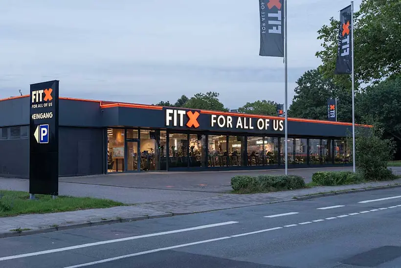 Bild: Außenansicht eines FitX Fitnessstudios. Ein großes FitX-Logo mit orangefarbenem X ist zu sehen. Das Gebäude befindet sich in einem urbanen Umfeld.