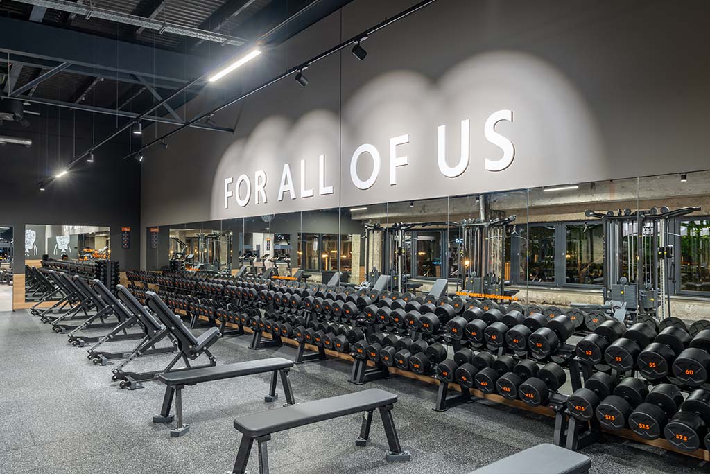 Weitläufiger Freihantelbereich mit verschiedenen Kraftgeräten im FitX Fitnessstudio Berlin-Ku'damm. An der Seite sind Kurzhanteln vor einer Spiegelwand aufgereiht. An der oberen Wand prangt der Schriftzug „FOR ALL OF US“.