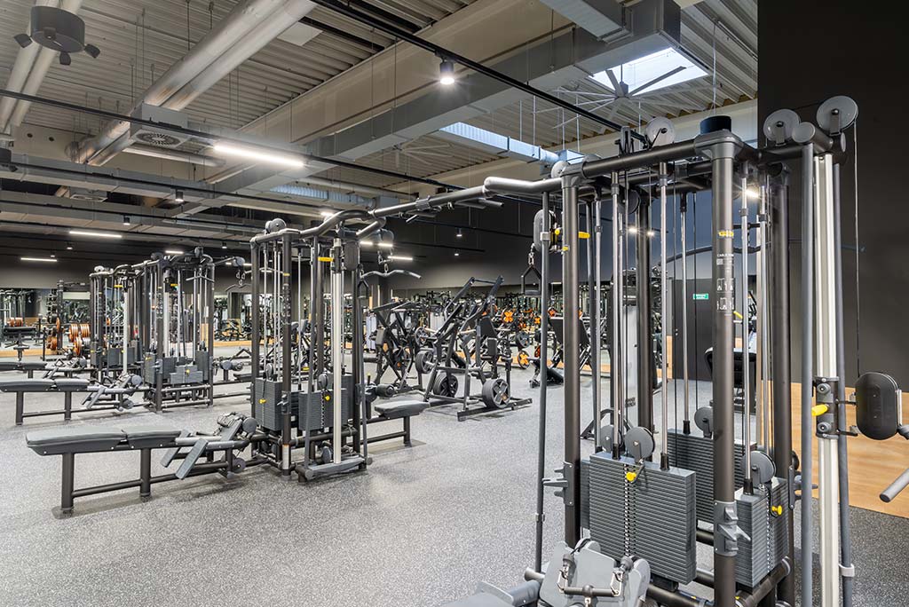 Weitläufiger Freihantelbereich mit verschiedenen Kraftgeräten im FitX Fitnessstudio Dortmund-Phoenix See. An der Seite sind Kurzhanteln vor einer Spiegelwand aufgereiht. An der oberen Wand prangt der Schriftzug „FOR ALL OF US“.
