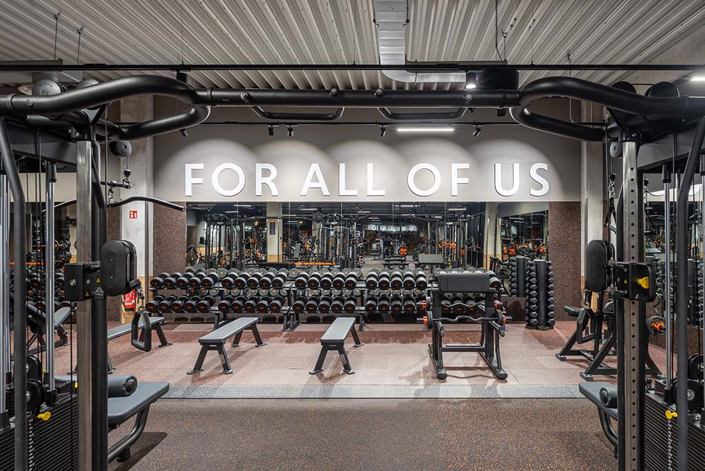 Weitläufiger Freihantelbereich mit verschiedenen Kraftgeräten im FitX Fitnessstudio Remscheid-Süd. An der Seite sind Kurzhanteln vor einer Spiegelwand aufgereiht. An der oberen Wand prangt der Schriftzug „FOR ALL OF US“.