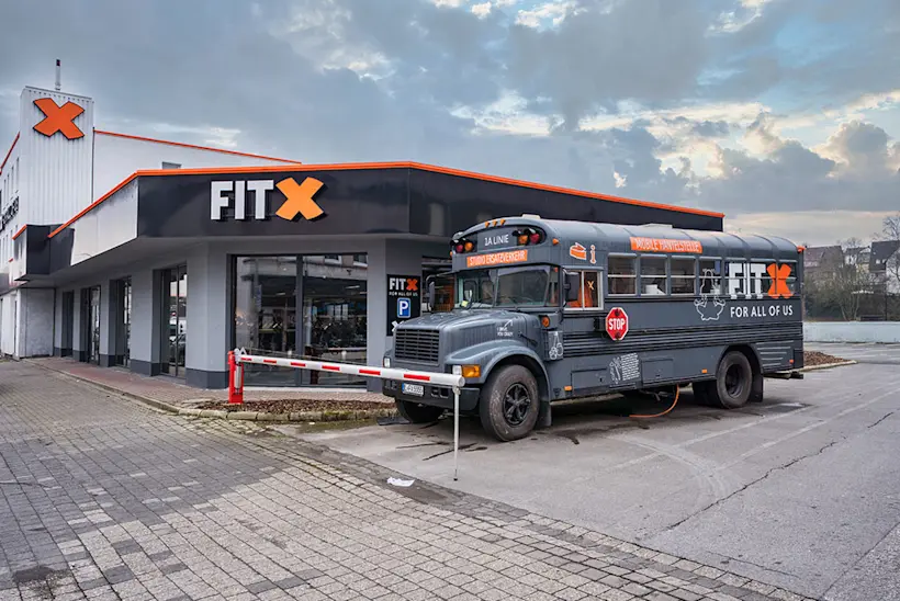 Bild: Außenansicht eines FitX Fitnessstudios. Ein großes FitX-Logo mit orangefarbenem X ist zu sehen. Das Gebäude befindet sich in einem urbanen Umfeld.