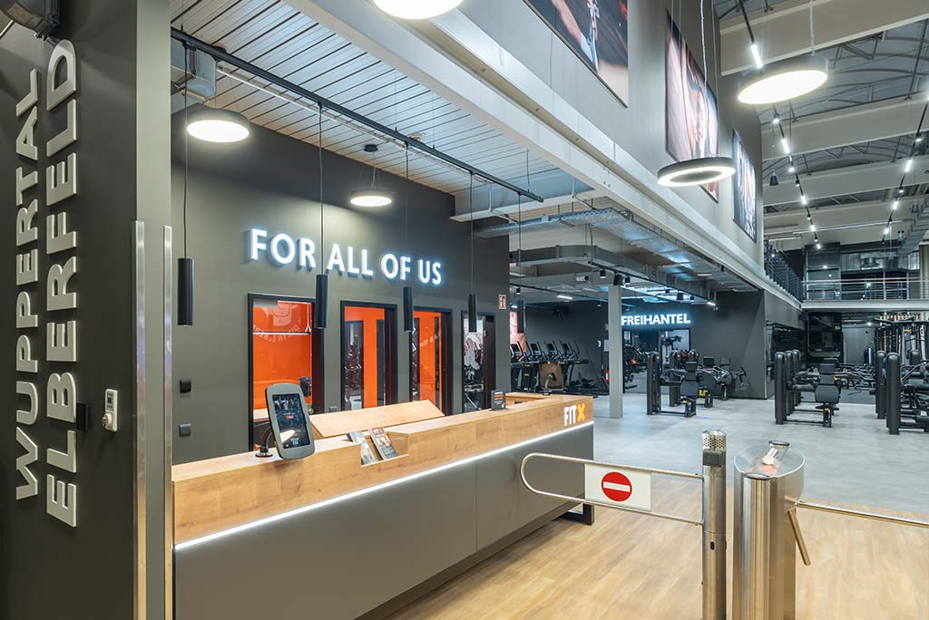 Empfangsbereich des FitX Fitnessstudios Wuppertal-Elberfeld mit einer holzverkleideten Theke, einem Drehkreuz am Eingang und dem leuchtenden Schriftzug „FOR ALL OF US“ an der Wand.