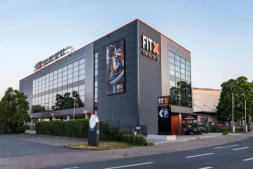 Bild: Außenansicht eines FitX Fitnessstudios. Ein großes FitX-Logo mit orangefarbenem X ist zu sehen. Das Gebäude befindet sich in einem urbanen Umfeld.