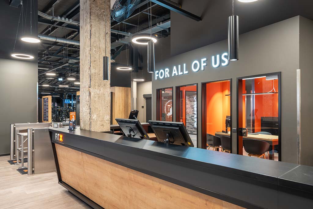 Empfangsbereich des FitX Fitnessstudios Berlin-Ku'damm mit einer holzverkleideten Theke, einem Drehkreuz am Eingang und dem leuchtenden Schriftzug „FOR ALL OF US“ an der Wand.
