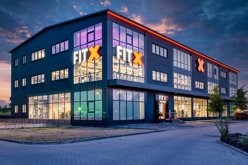 Außenansicht des FitX Fitnessstudios Leipzig-Neulindenau. Ein großes FitX-Logo mit orangefarbenem X ist zu sehen. Das Gebäude befindet sich in einem urbanen Umfeld.