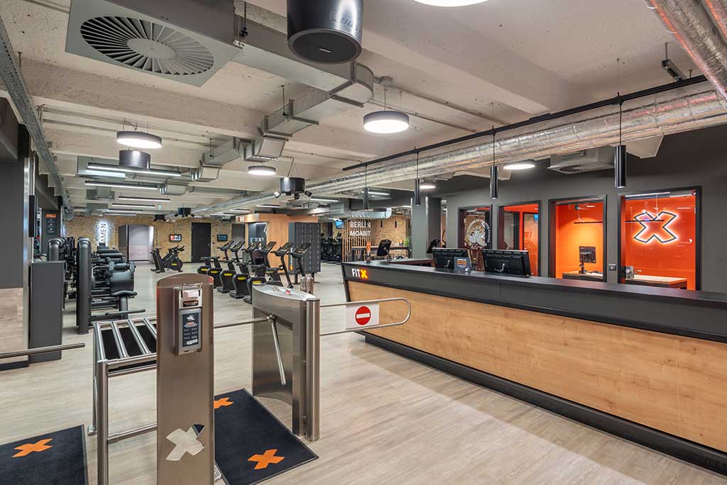 Empfangsbereich des FitX Fitnessstudios Berlin-Moabit mit einer holzverkleideten Theke, einem Drehkreuz am Eingang und dem leuchtenden Schriftzug „FOR ALL OF US“ an der Wand.