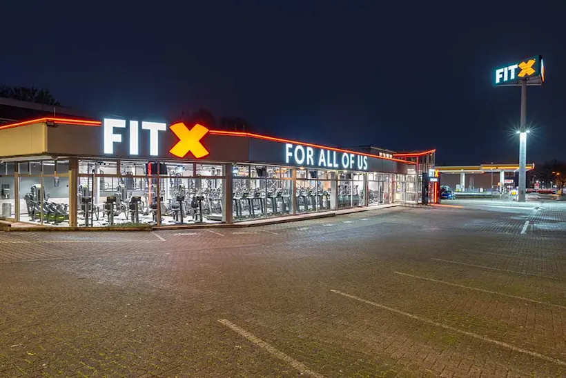 Bild: Außenansicht eines FitX Fitnessstudios. Ein großes FitX-Logo mit orangefarbenem X ist zu sehen. Das Gebäude befindet sich in einem urbanen Umfeld.