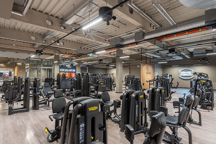 Weitläufiger Freihantelbereich mit verschiedenen Kraftgeräten im FitX Fitnessstudio Augsburg-Lechhausen. An der Seite sind Kurzhanteln vor einer Spiegelwand aufgereiht. An der oberen Wand prangt der Schriftzug „FOR ALL OF US“.
