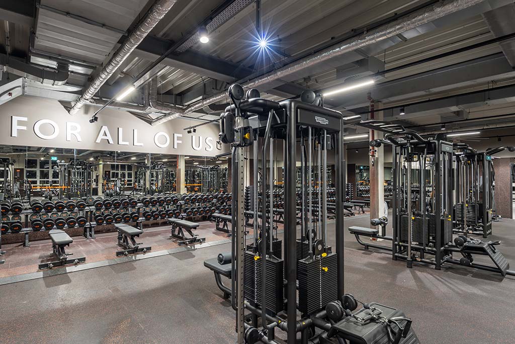 Weitläufiger Freihantelbereich mit verschiedenen Kraftgeräten im FitX Fitnessstudio Mülheim-Stadtmitte. An der Seite sind Kurzhanteln vor einer Spiegelwand aufgereiht. An der oberen Wand prangt der Schriftzug „FOR ALL OF US“.