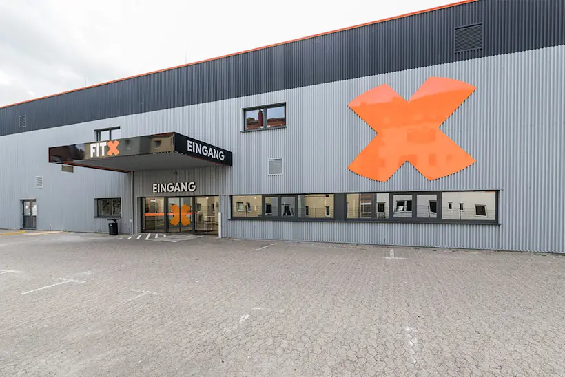 Bild: Außenansicht eines FitX Fitnessstudios. Ein großes FitX-Logo mit orangefarbenem X ist zu sehen. Das Gebäude befindet sich in einem urbanen Umfeld.