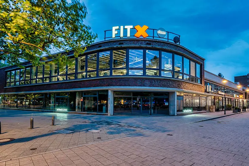 Bild: Außenansicht eines FitX Fitnessstudios. Ein großes FitX-Logo mit orangefarbenem X ist zu sehen. Das Gebäude befindet sich in einem urbanen Umfeld.