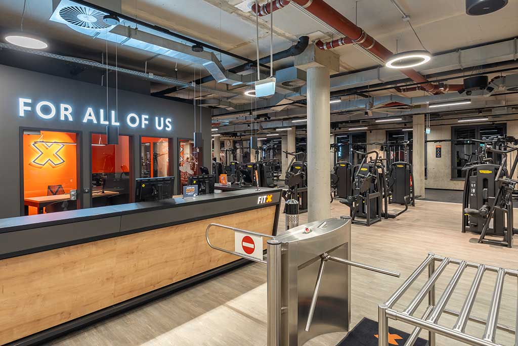 Empfangsbereich des FitX Fitnessstudios Troisdorf-Mitte mit einer holzverkleideten Theke, einem Drehkreuz am Eingang und dem leuchtenden Schriftzug „FOR ALL OF US“ an der Wand.