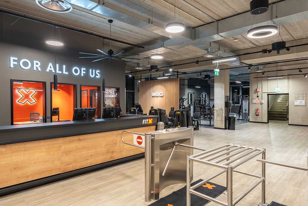 Empfangsbereich des FitX Fitnessstudios Leipzig-Neulindenau mit einer holzverkleideten Theke, einem Drehkreuz am Eingang und dem leuchtenden Schriftzug „FOR ALL OF US“ an der Wand.