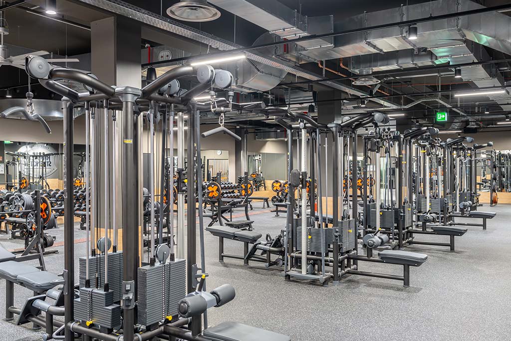 Weitläufiger Freihantelbereich mit verschiedenen Kraftgeräten im FitX Fitnessstudio Aachen-Mitte. An der Seite sind Kurzhanteln vor einer Spiegelwand aufgereiht. An der oberen Wand prangt der Schriftzug „FOR ALL OF US“.