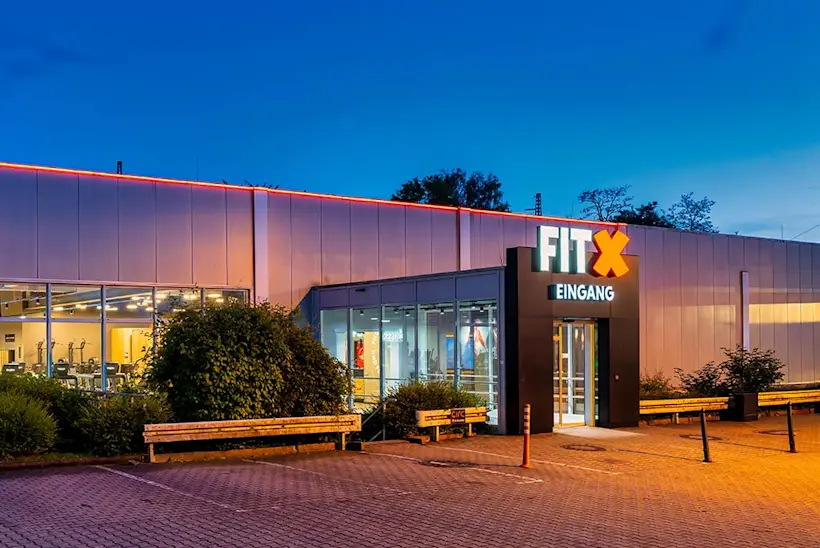 Bild: Außenansicht eines FitX Fitnessstudios. Ein großes FitX-Logo mit orangefarbenem X ist zu sehen. Das Gebäude befindet sich in einem urbanen Umfeld.