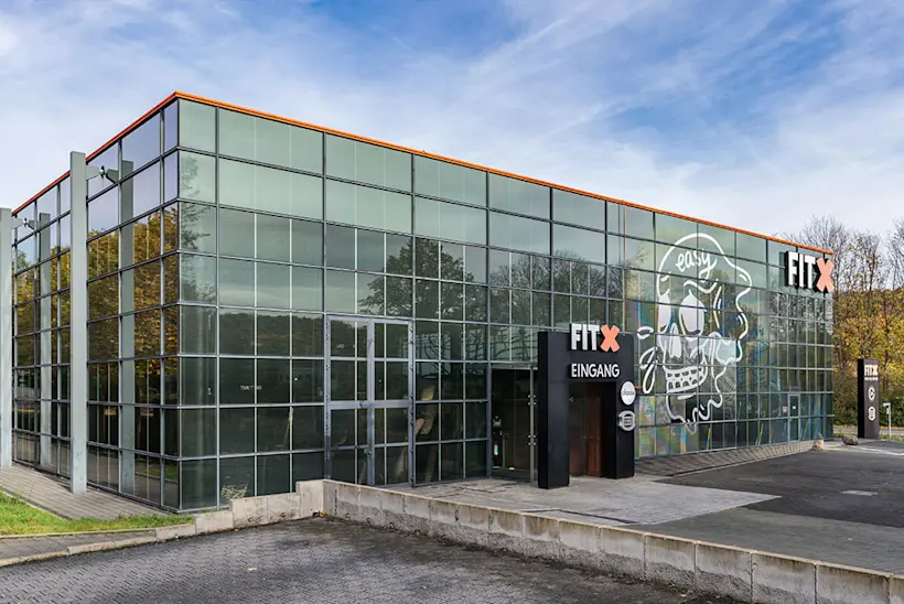 Bild: Außenansicht eines FitX Fitnessstudios. Ein großes FitX-Logo mit orangefarbenem X ist zu sehen. Das Gebäude befindet sich in einem urbanen Umfeld.