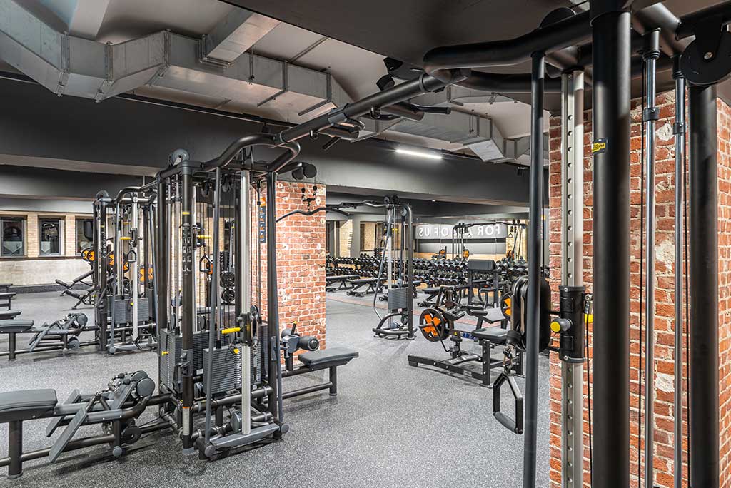 Weitläufiger Freihantelbereich mit verschiedenen Kraftgeräten im FitX Fitnessstudio Berlin-Moabit. An der Seite sind Kurzhanteln vor einer Spiegelwand aufgereiht. An der oberen Wand prangt der Schriftzug „FOR ALL OF US“.