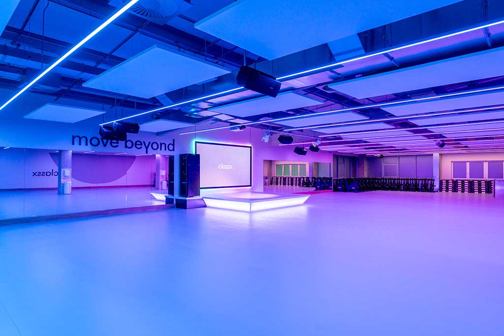 Großer, offener Kursraum im FitX Fitnessstudio Leipzig-Neulindenau. An der hinteren Wand befindet sich eine erhöhte Bühne, dahinter eine Leinwand. Links und rechts davon sind Spiegelwände.