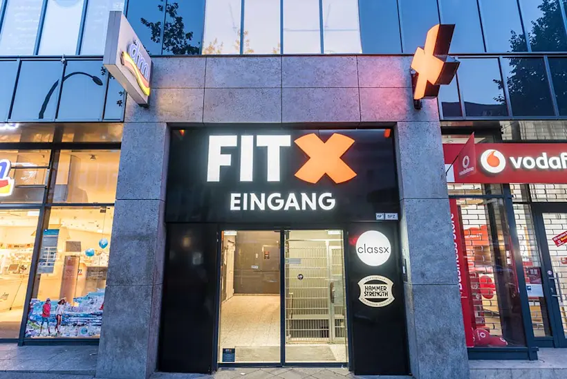 Bild: Außenansicht eines FitX Fitnessstudios. Ein großes FitX-Logo mit orangefarbenem X ist zu sehen. Das Gebäude befindet sich in einem urbanen Umfeld.