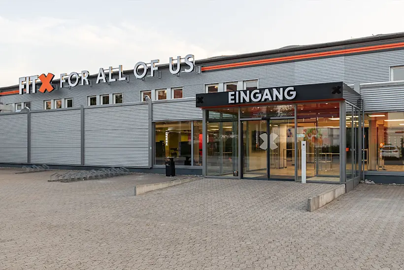 Bild: Außenansicht eines FitX Fitnessstudios. Ein großes FitX-Logo mit orangefarbenem X ist zu sehen. Das Gebäude befindet sich in einem urbanen Umfeld.