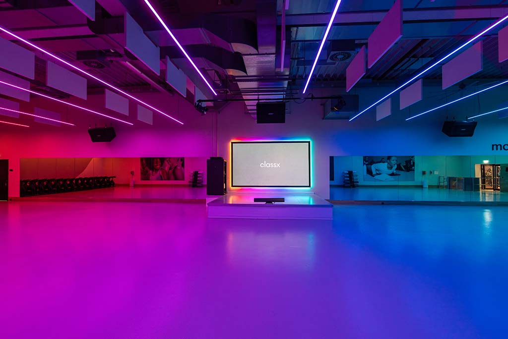 Großer, offener Kursraum im FitX Fitnessstudio Dortmund-Phoenix See. An der hinteren Wand befindet sich eine erhöhte Bühne, dahinter eine Leinwand. Links und rechts davon sind Spiegelwände.