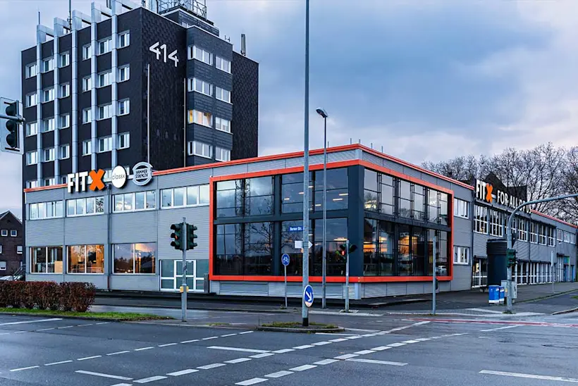Bild: Außenansicht eines FitX Fitnessstudios. Ein großes FitX-Logo mit orangefarbenem X ist zu sehen. Das Gebäude befindet sich in einem urbanen Umfeld.