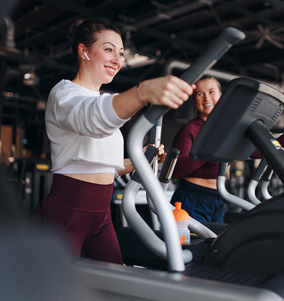 Bild: Zwei junge Frauen trainieren nebeneinander auf Crosstrainern im Cardiobereich. Beide lächeln und tragen Sportkleidung.