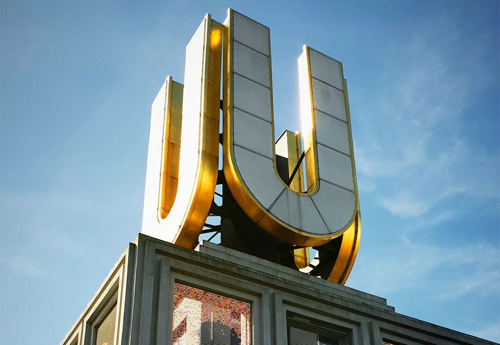 Bild: Nahaufnahme des Dortmunder U, einem hohen Gebäude mit einem großen, goldumrandeten „U“ auf dem Dach vor blauem Himmel.