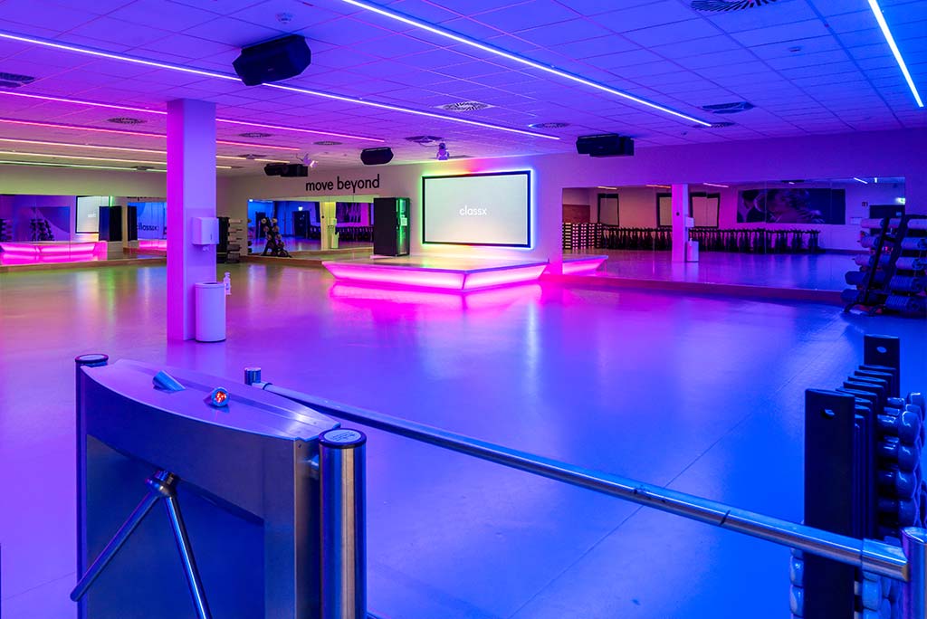 Großer, offener Kursraum im FitX Fitnessstudio Augsburg-Lechhausen. An der hinteren Wand befindet sich eine erhöhte Bühne, dahinter eine Leinwand. Links und rechts davon sind Spiegelwände.